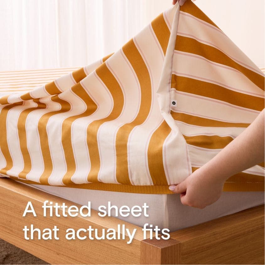 Close-up of a hand stretching out a fitted sheet with visible elastic corners. Overlay text reads 'A fitted sheet that actually fits.